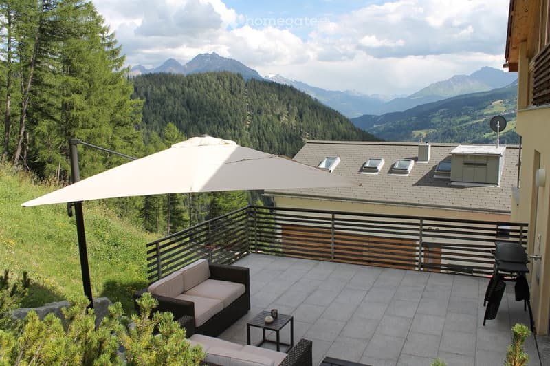 Balcony with a view of mountains, featuring a sofa, small table, umbrella, and grill.