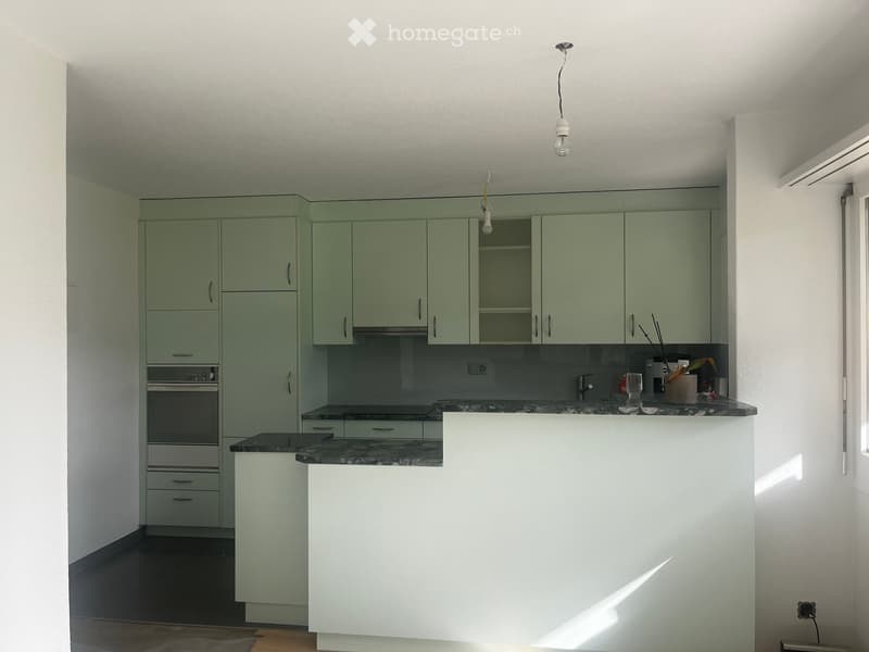 Modern kitchen with light green cabinets, dark countertops, and built-in oven. Features a central island with a white surface and a hanging light fixture.
