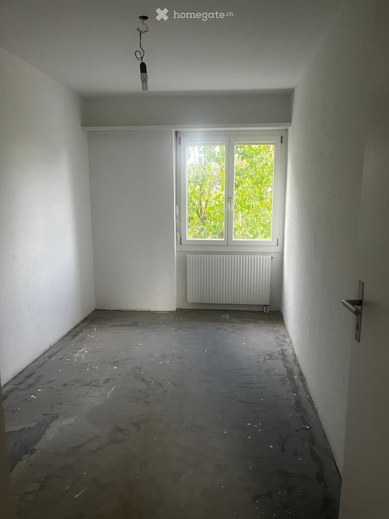 Empty room with white walls, concrete floor, a single window with a radiator below, and a hanging light fixture.