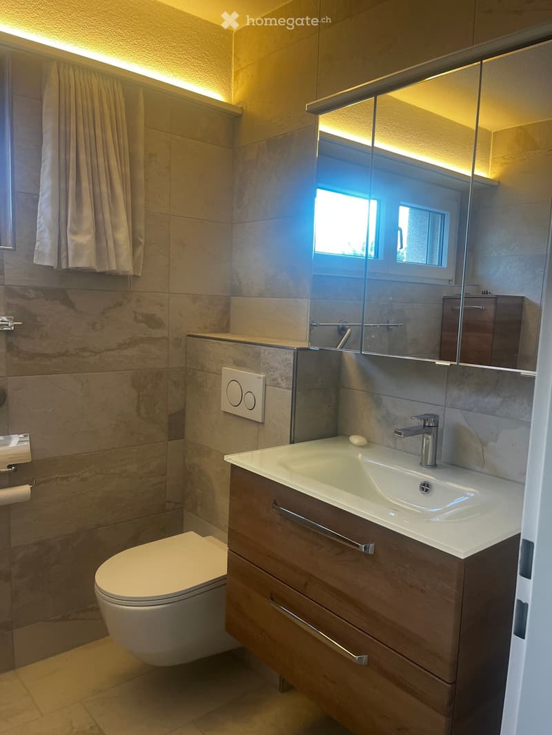 Modern bathroom with beige tiles, a white toilet, a wooden vanity with a sink and faucet, a large mirror, and a towel hanging on the wall.