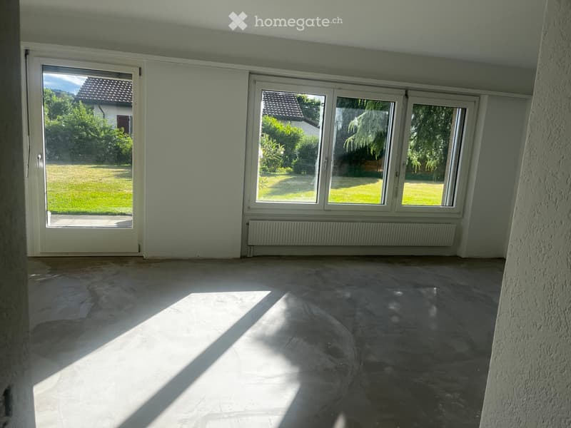 Empty room with large windows and sliding doors leading to a garden, featuring a tiled floor and light-colored walls.