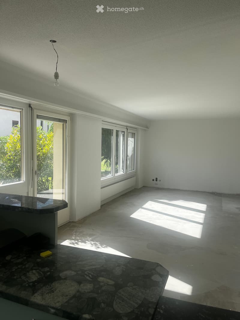 Empty room with white walls, large windows, and a stone countertop. Sunlight streams in, casting shadows on the floor.