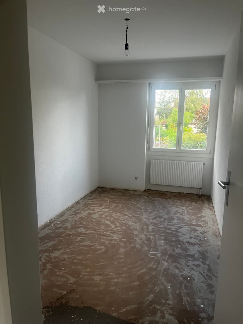 Empty room with white walls, a single window with white frame, a radiator, and a light fixture on the ceiling. The floor is unfinished and shows exposed concrete and wood.