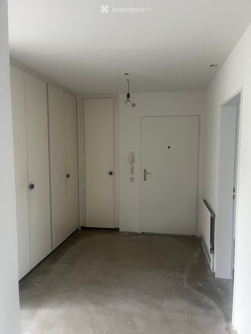 Empty hallway with white walls, concrete floor, and several doors. A light fixture hangs from the ceiling, and a telephone is mounted on one wall.