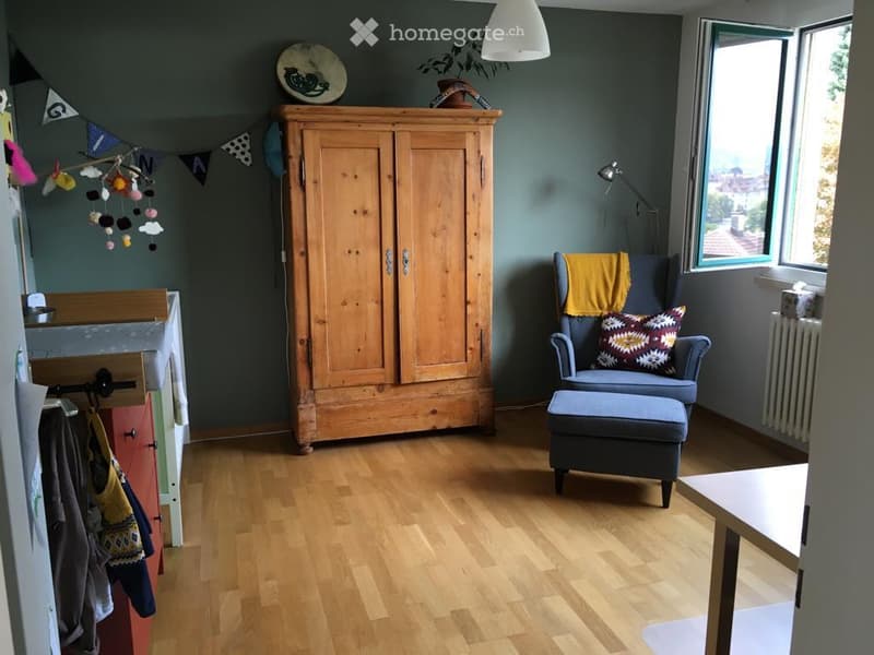 A cozy, well-lit room with wooden flooring, a large wooden wardrobe, a blue armchair with a patterned cushion and matching ottoman, a small table, and a decorative plate on top of the wardrobe. The room also features a hanging toy mobile and a coat rack w