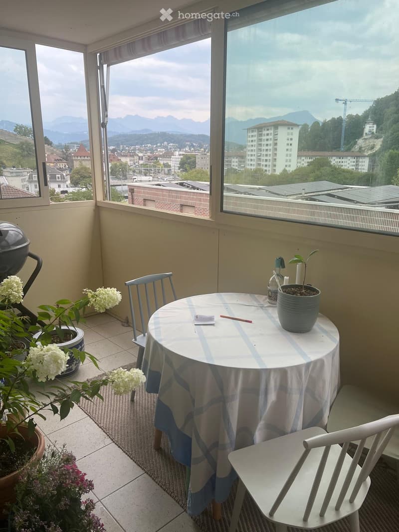 Balcony with a round table, two chairs, potted plants, and a scenic city view with mountains in the background.