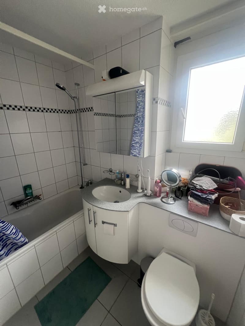 Bathroom with a bathtub, sink, toilet, mirror, and various toiletries on the counter. The room features tiled walls and a window letting in natural light.