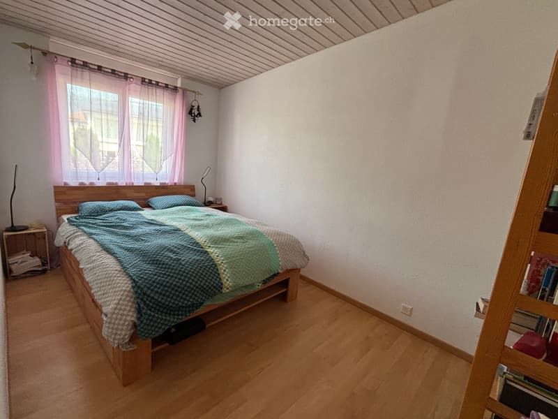 Cozy bedroom with wooden floor, large window with pink curtains, bed with blue and green quilt, nightstand with lamp, and bookshelf with books.
