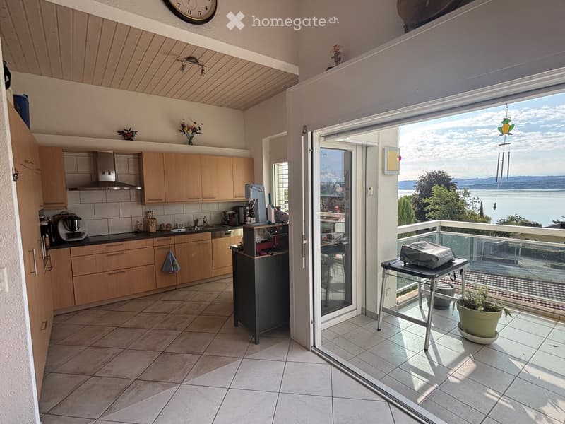 Modern kitchen with light wooden cabinets, stainless steel appliances, and a large window opening to a balcony with a scenic view of a lake.
