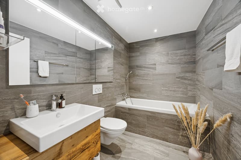 Modern bathroom with grey tiled walls, a white sink with wooden cabinet, a toilet, a bathtub, a large mirror, and a vase with dried plants on the floor.