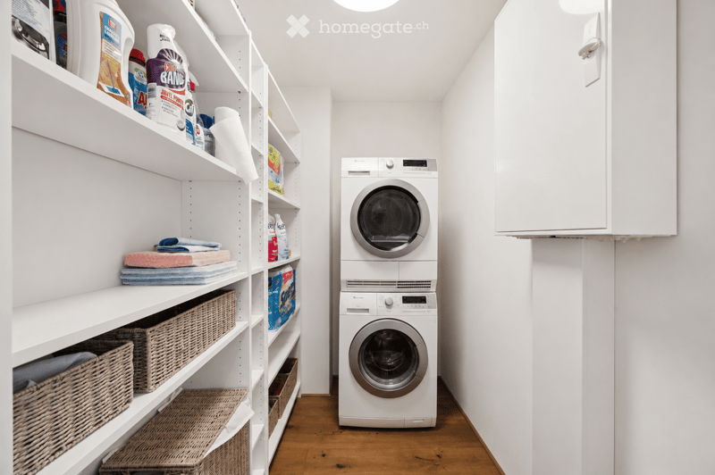 A modern laundry room with white walls, wooden flooring, and built-in white shelving units holding cleaning supplies, towels, and wicker baskets. A stacked washer and dryer unit is positioned in the center.