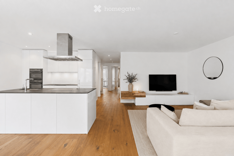 Open-plan living and dining area with a modern kitchen, white cabinets, and a central island. The living area features a light-colored sofa, a coffee table, and a flat-screen TV mounted on the wall. The space has wooden flooring and large windows allowing