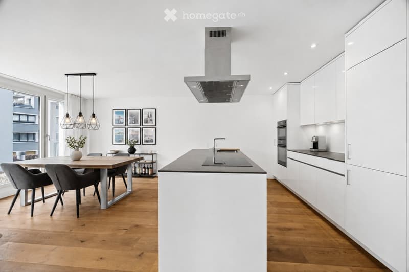Modern kitchen with white cabinets, dark countertops, a central island, and a dining area with a wooden table and grey chairs. Features pendant lights, a range hood, and a coffee machine on the counter.