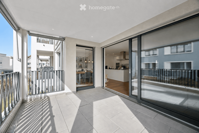 Modern balcony with large glass sliding doors, tiled floor, metal railing, and views of neighboring buildings.
