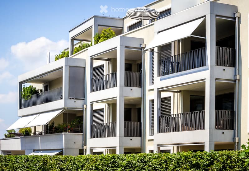 Modern multi-story residential building with balconies, large windows, and greenery on the roof and balconies.