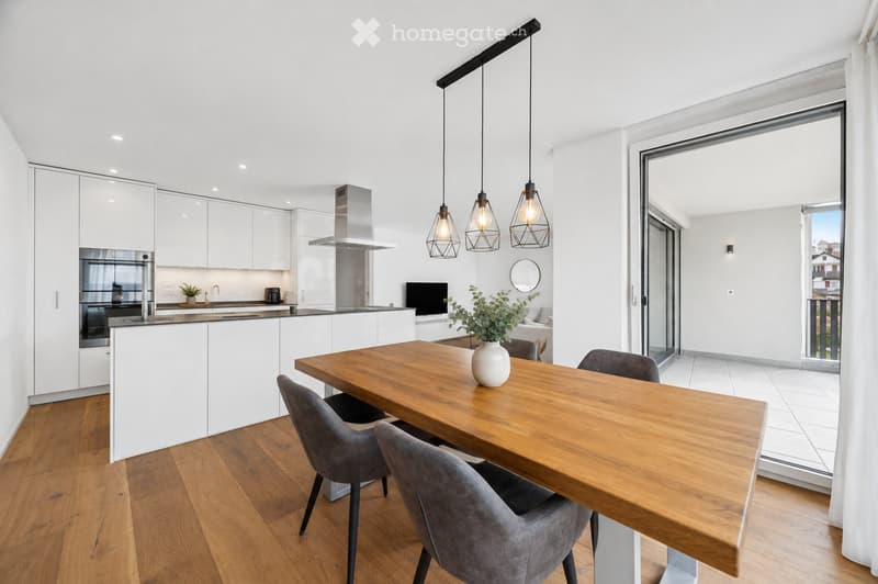 Modern kitchen with white cabinets, wooden dining table, pendant lights, and a view to a balcony.
