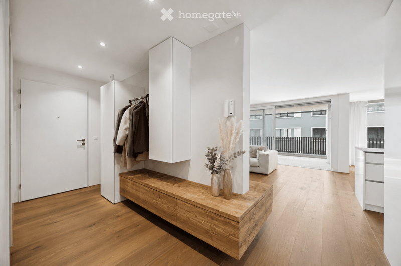 Modern hallway with wooden flooring, white walls, a built-in bench with vases, a coat rack, and a view to a living area with a sofa and balcony.
