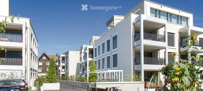 Modern multi-story residential building with white facade, large windows, and balconies. The building is surrounded by greenery and trees, with a parking area in the foreground.