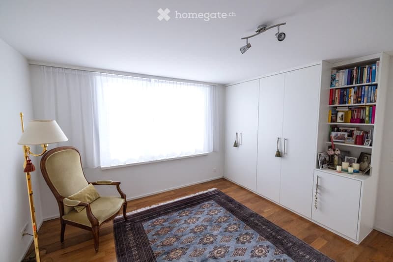 Bright room with wooden flooring, large window with white curtains, a single armchair with a cushion, a bookshelf filled with books and decorative items, a table lamp, and a patterned rug.