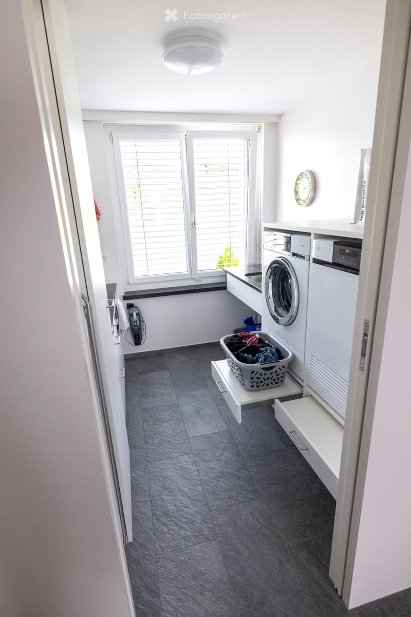 Bright, modern bathroom with grey tiled floor, white walls, and built-in cabinets. Features a washing machine, dishwasher, laundry basket, and a clock on the wall.