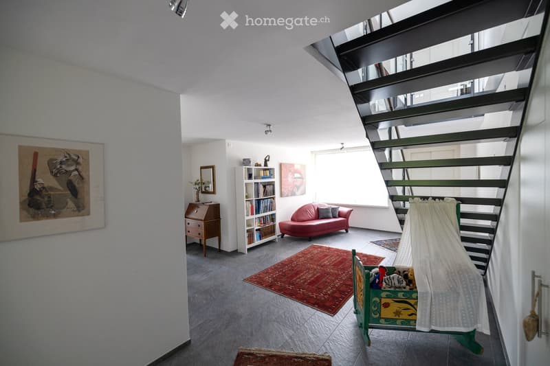 Bright living room with a red sofa, bookshelf, small table, and artwork on the walls. A staircase leads upwards, and a toy box with a canopy is visible near the stairs.