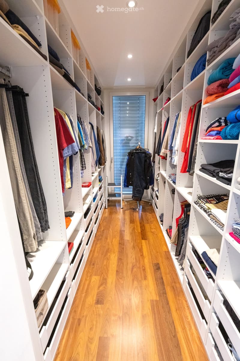 Spacious, well-lit hallway with built-in white shelving units on both sides, filled with neatly organized clothing and folded items. The floor is made of light wooden planks, and the ceiling features recessed lighting.