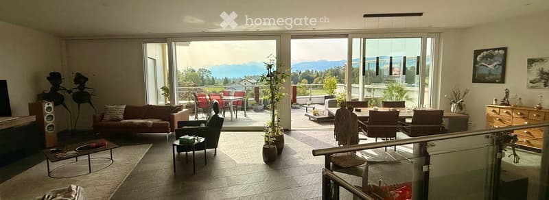 Modern living room with large glass doors opening to a balcony, offering a scenic mountain view. The room features a brown sofa, coffee table, armchair, fireplace, dining table with chairs, and several potted plants. A sleek speaker system and a wooden ca