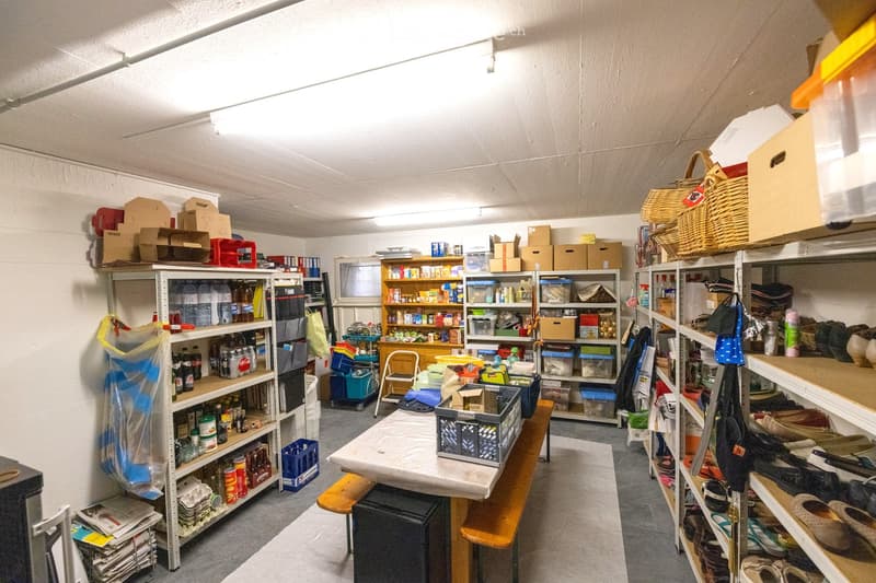 Storage room with multiple shelves filled with various items including bottled drinks, plastic containers, clothing, shoes, and kitchen supplies. A wooden bench with a table on top holds additional storage items.