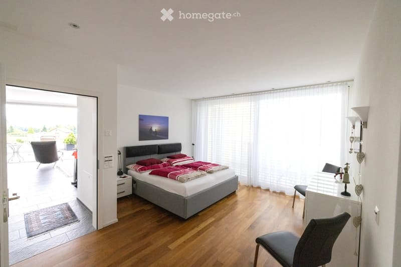 Bright bedroom with wooden flooring, large bed with red and white bedding, nightstand, chair, table, and a view to a balcony area.