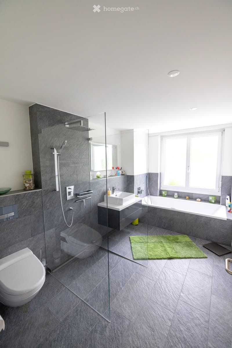 Modern bathroom with grey tiles, glass shower enclosure, toilet, bathtub, sink, and a green rug on the floor.