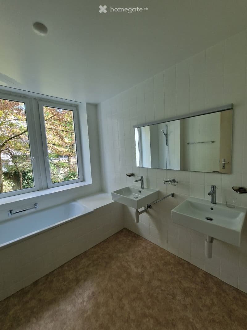 Modern bathroom with a bathtub, two sinks, a large mirror, and a window offering a view of greenery.