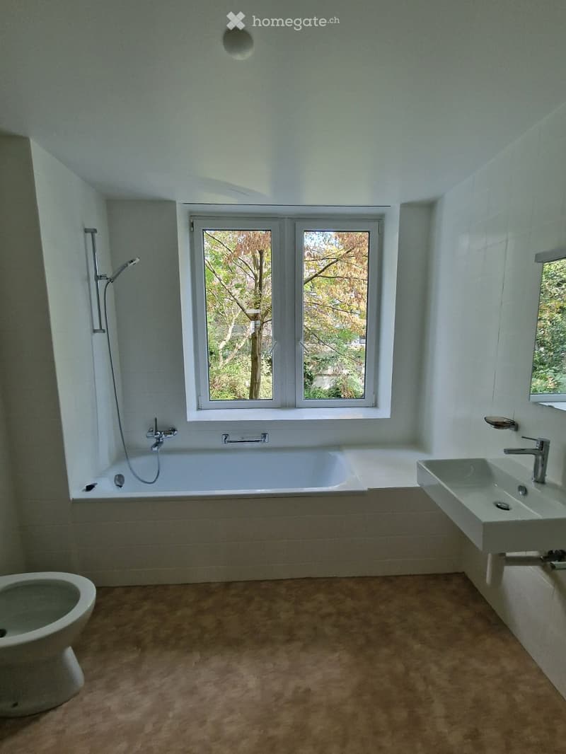 Modern bathroom with a bathtub, toilet, sink, and showerhead. Large window provides natural light, and the space features light-colored tiles and a warm-toned floor.