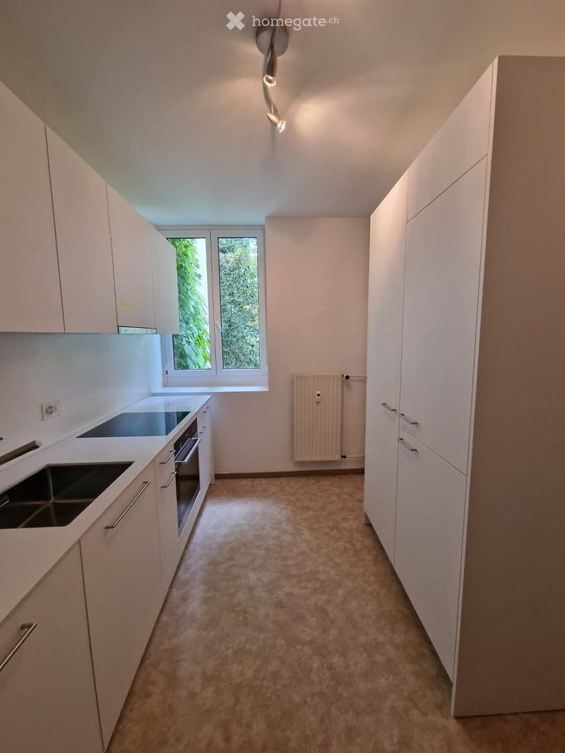Modern kitchen with white cabinets, stainless steel sink, and oven. Light-colored carpet flooring and large windows provide natural light.
