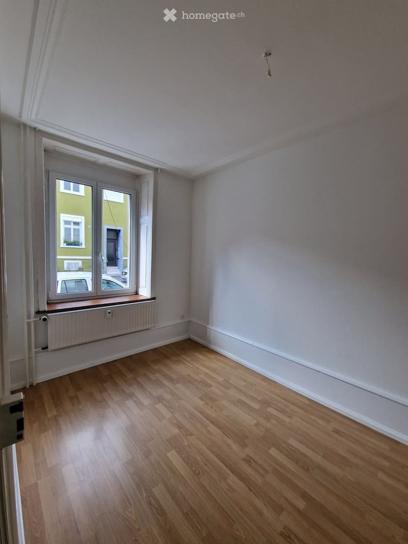 Empty room with light wooden flooring, white walls, and a large window with a radiator below it.