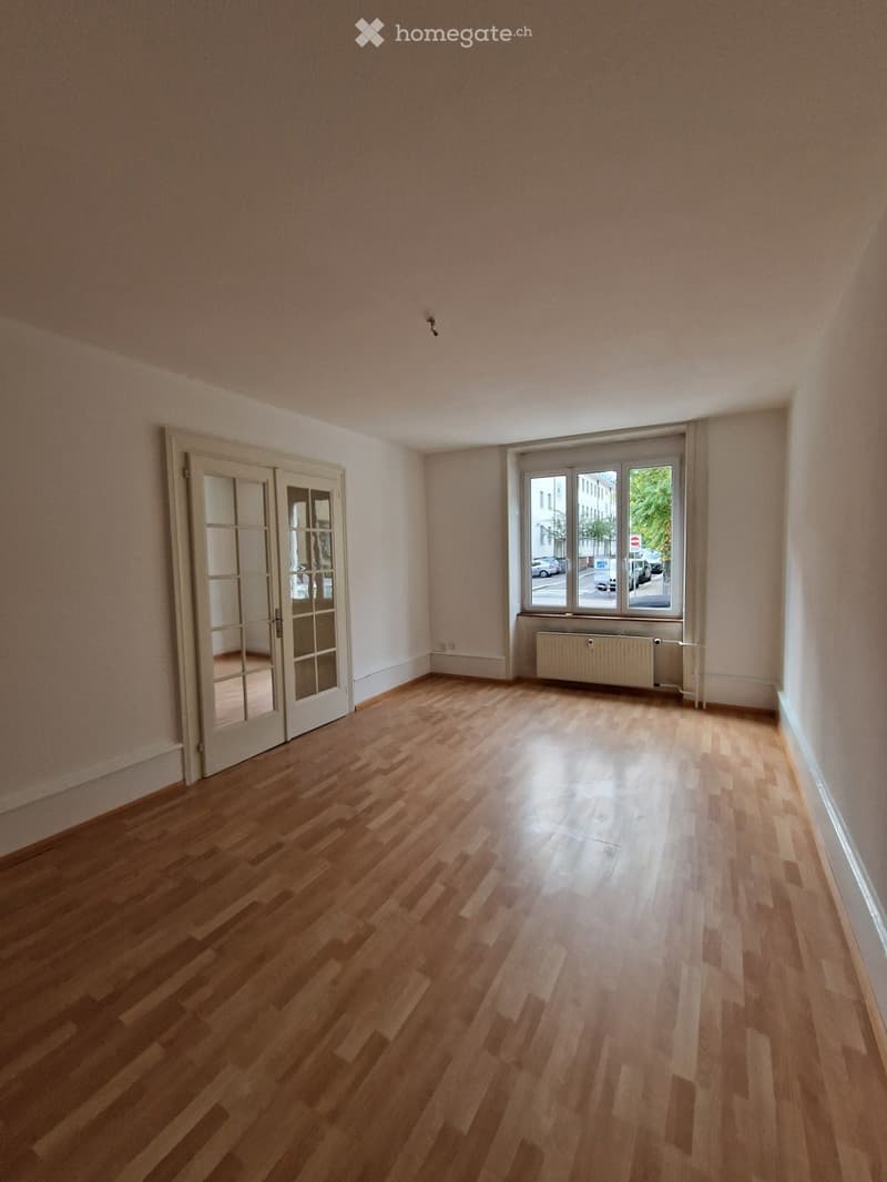 Empty room with wooden flooring, large window, and double doors leading to another space.