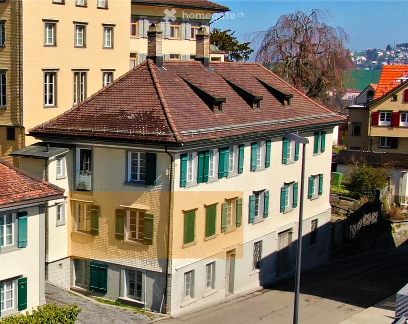 A multi-story residential building with a red-tiled roof, green shutters, and multiple windows. The building is part of a row of similar structures in a residential area.
