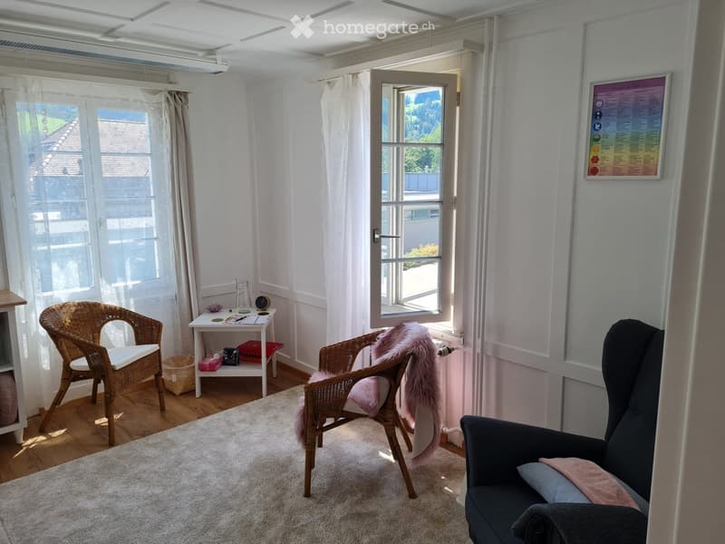 Bright living room with white walls, wooden floor, large windows with sheer curtains, a sofa, wicker chairs, a small table, and a rug.