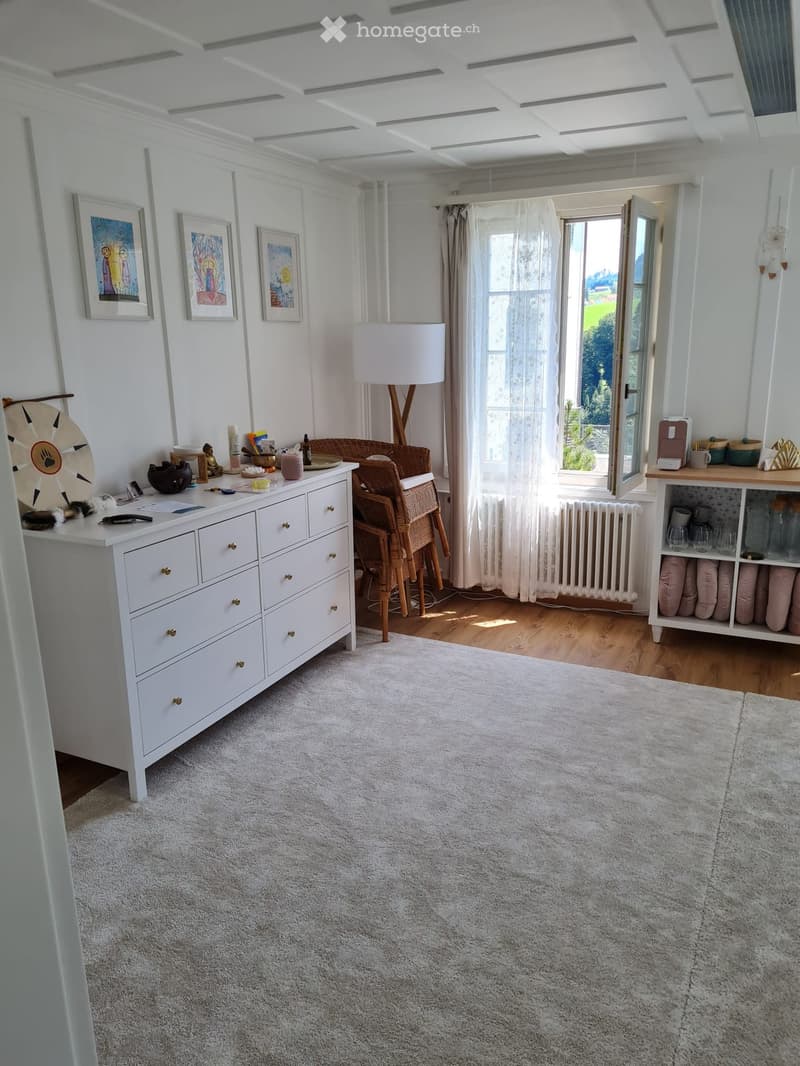 Bright, airy room with white walls, wooden floor, large window with sheer curtains, and a view of greenery. Features a white dresser, a wicker chair, a floor lamp, and a shelf with decorative items.