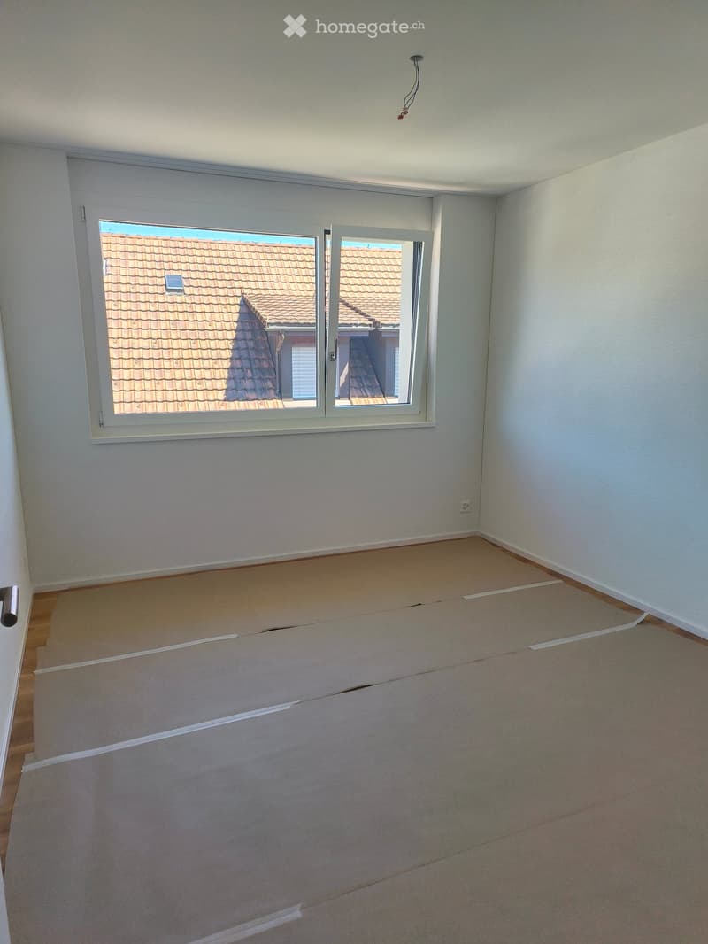 Empty room with light-colored walls, wooden flooring, and a large window showing a tiled roof outside. No furniture or equipment present.