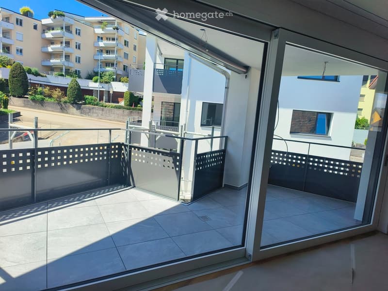 Balcony with tiled floor, glass sliding doors, metal railing, and view of surrounding buildings and greenery.