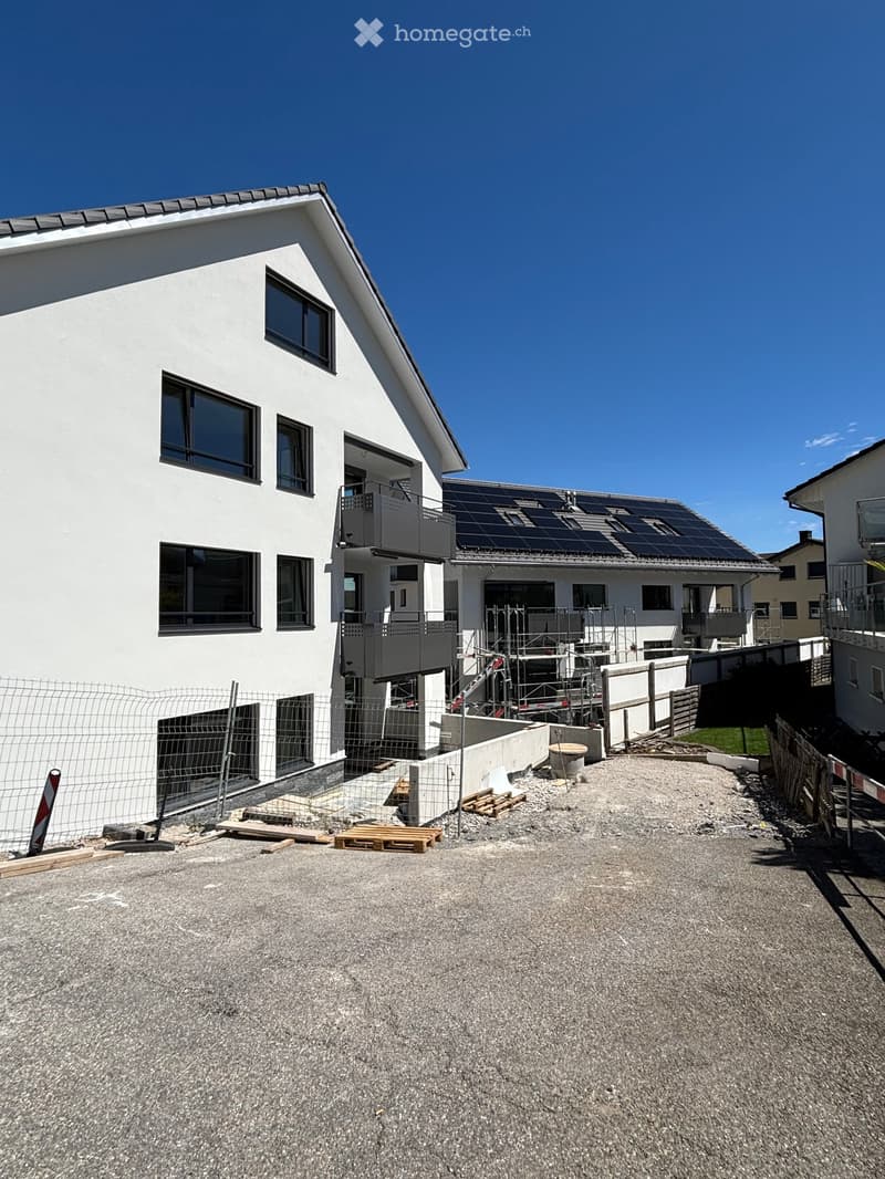 Modern two-story house with white exterior, large windows, solar panels on the roof, and a fenced construction area with pallets and materials.