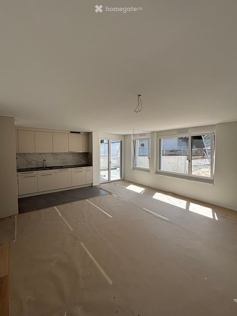 Empty room with light-colored walls, large windows, and a small kitchen area with cabinets and a sink.