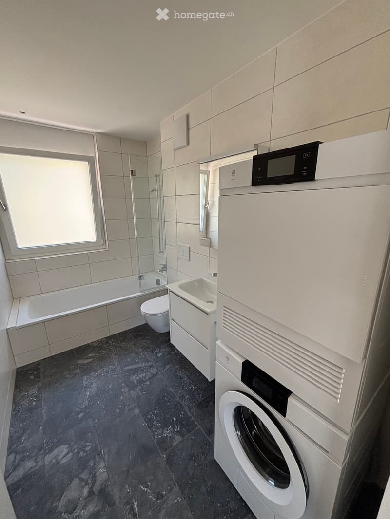 Modern bathroom with dark marble flooring, white tiled walls, bathtub, toilet, sink, and a stacked washing machine and dryer unit.