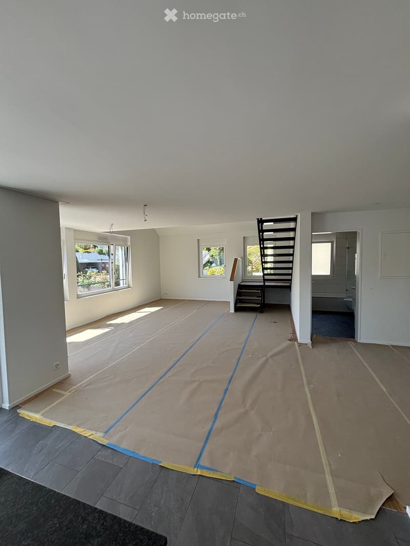 Empty room with bare walls and floor, covered with protective sheets and tape, with visible windows and a staircase leading to another area.