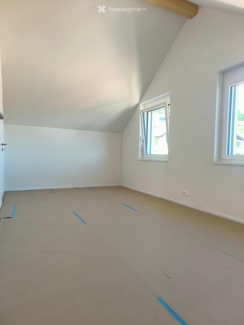 Empty attic room with white walls, slanted ceiling, and two windows, one of which is open. Blue painter's tape marks the floor.