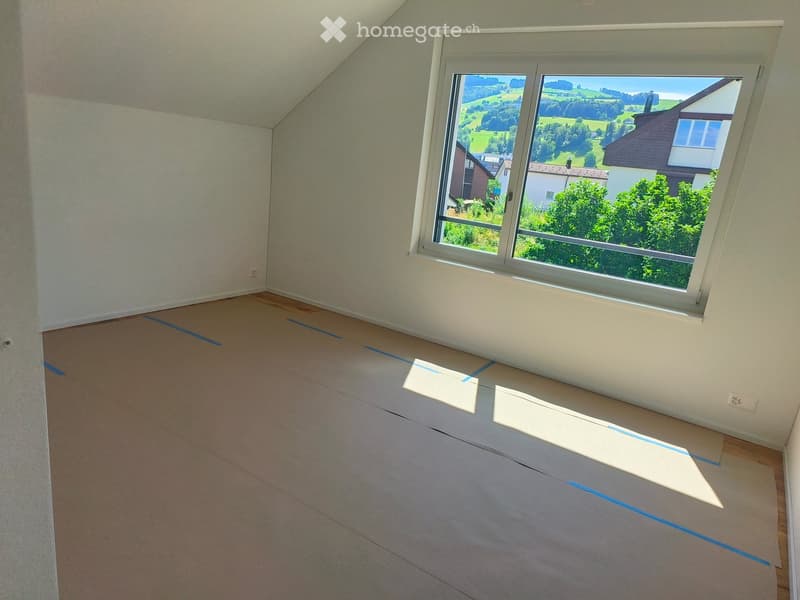 Empty attic room with wooden floor and large window showing a scenic view of green fields and houses. Blue painter's tape marks the floor.