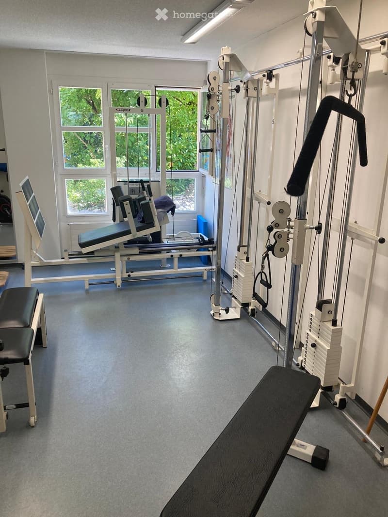 Well-lit gym room with various exercise equipment including a bench, weight machine, cable machine, weight stack, and a stool.