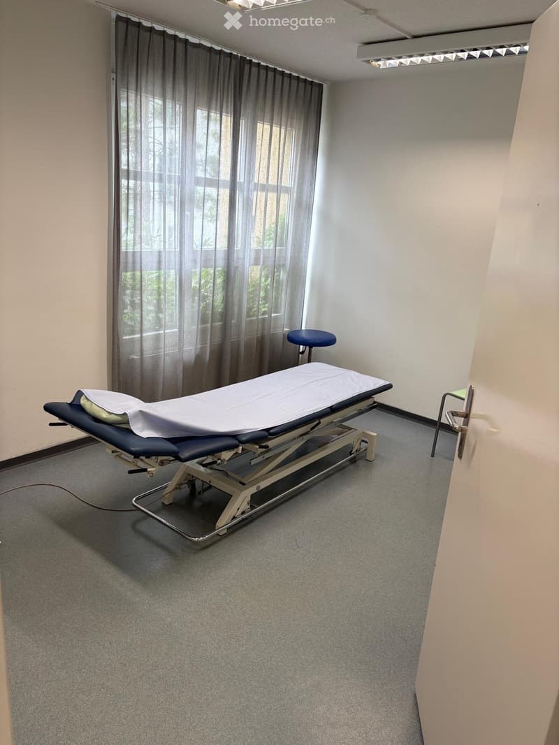 A clean, well-lit room with a treatment table and a stool, featuring large windows with curtains and a grey carpeted floor.