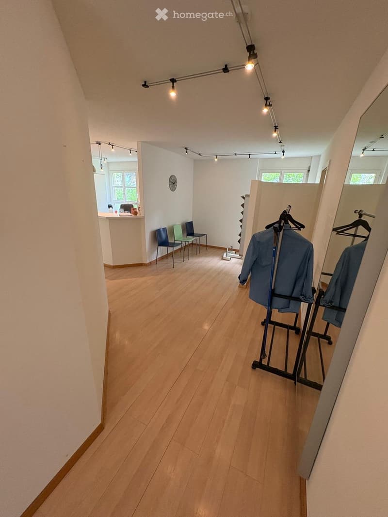 A well-lit hallway with wooden flooring, featuring a clothing rack with jackets, a mirror, and a small seating area with chairs.