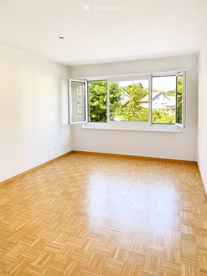 Empty room with light wooden herringbone flooring and white walls, featuring large windows with open blinds showing greenery outside.